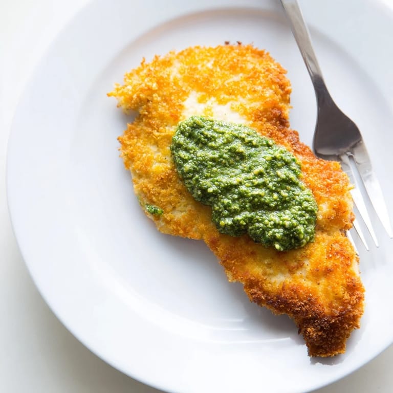 Golden, crunchy Parmesan breadcrumbs top juicy chicken cutlets, finished with a vibrant green dollop of basil pesto on a platter.