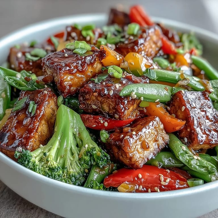 A close-up of Vegetable Tempeh Stir-Fry reveals colorful bell peppers, snap peas, and broccoli coated in a glossy, aromatic ginger-soy glaze.